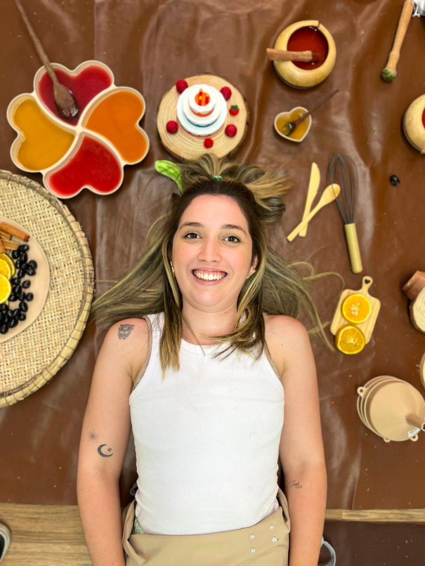 Bruna Aranda deitada no chão da oficina, entre materiais do brincar, também como mãe.